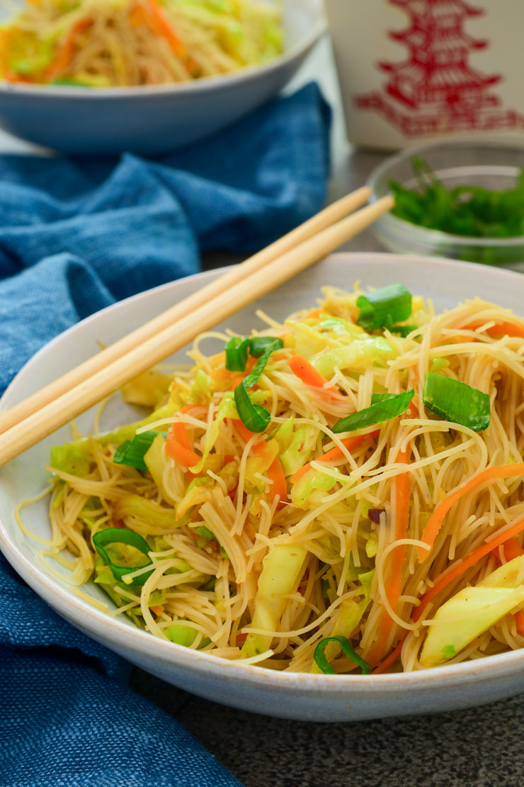 A white bowl of rice vermicelli and stir fried vegetables with chopsticks on the side.