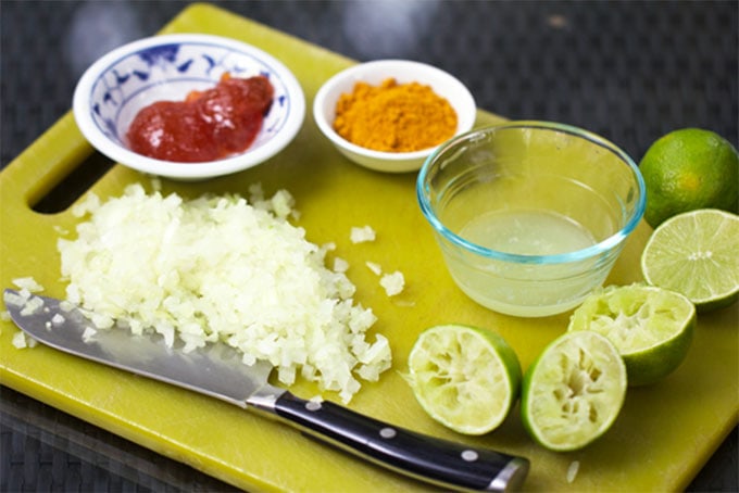 green cutting board topped with chopped onions, halved juiced limes, a small bowl of mango chutney, a small bowl of curry powder and a small bowl of lime juice